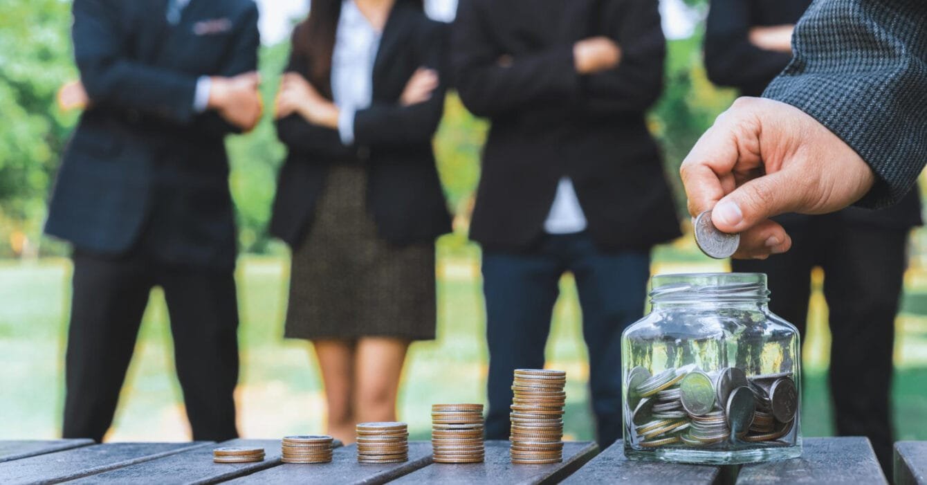 A person placing a coin into a jar with stacked coins on a table, businesspeople in the background, representing Public-Private Partnerships in Cambodia Constructionv.