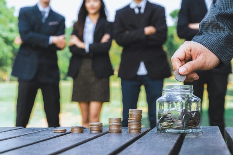 A person placing a coin into a jar with stacked coins on a table, businesspeople in the background, representing Public-Private Partnerships in Cambodia Constructionv.