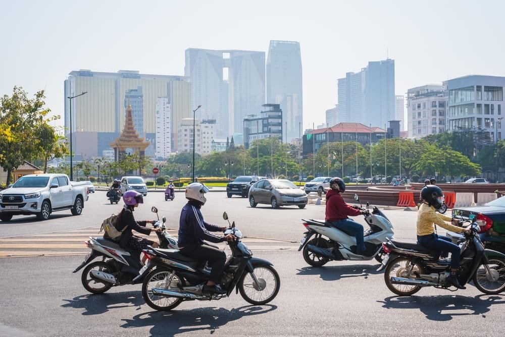 Motorcyclists on Phnom Penh city road with cars and modern skyscrapers in the background, showing the needs of Affordable Housing in Cambodia.