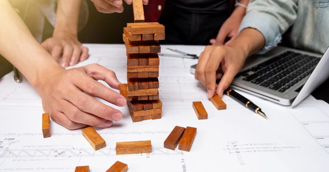 Cambodia Construction Industry Challenges are symbolised by hands playing Jenga on blueprint-covered desk with laptop nearby.