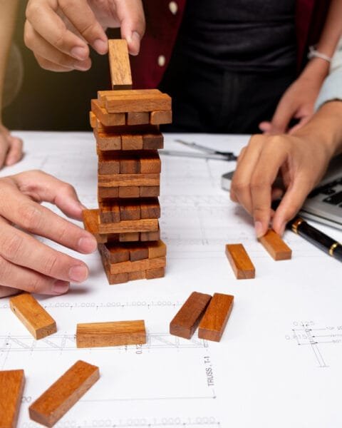 Cambodia Construction Industry Challenges are symbolised by hands playing Jenga on blueprint-covered desk with laptop nearby.