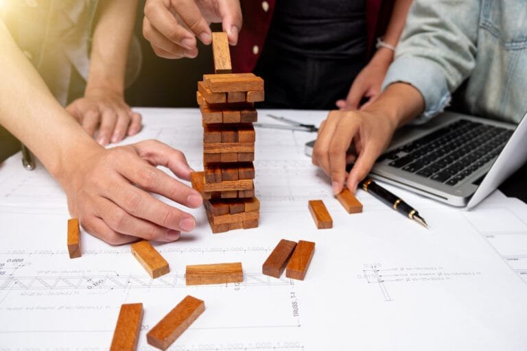 Cambodia Construction Industry Challenges are symbolised by hands playing Jenga on blueprint-covered desk with laptop nearby.