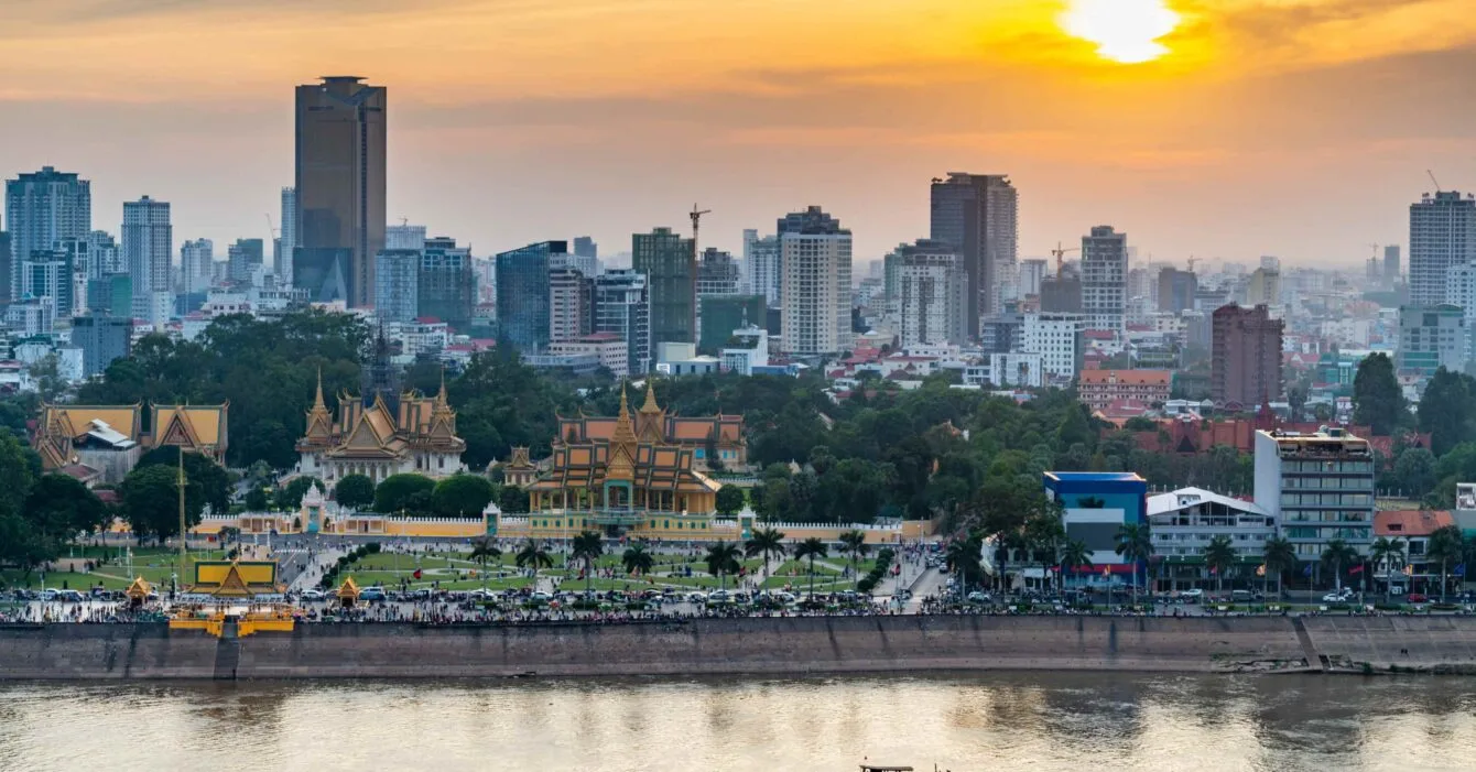 Cambodia Infrastructure Investments: Sunset over a river with traditional buildings in the foreground and modern cityscape in the background.