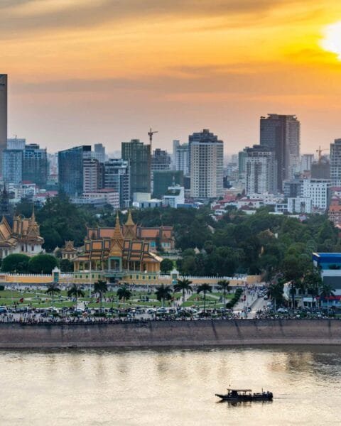 Cambodia Infrastructure Investments: Sunset over a river with traditional buildings in the foreground and modern cityscape in the background.