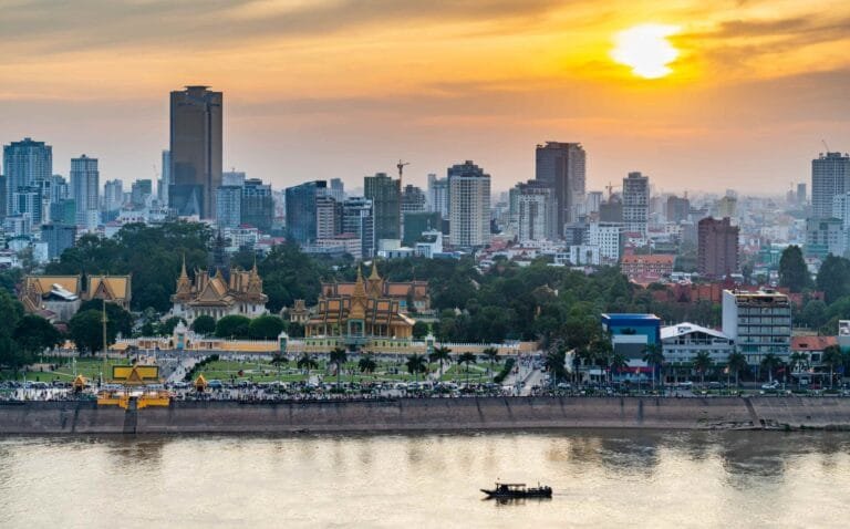 Cambodia Infrastructure Investments: Sunset over a river with traditional buildings in the foreground and modern cityscape in the background.