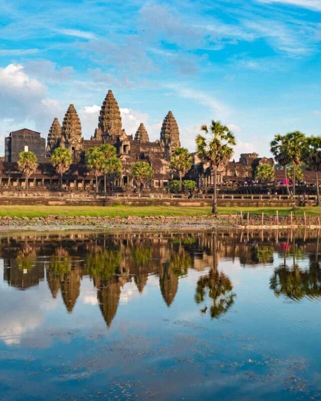 Angkor Wat temple complex reflected in a serene pond, surrounded by palm trees and lush greenery under a bright blue sky to represent Cambodia Tourism Industry Recovery.