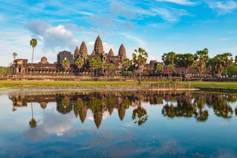 Angkor Wat temple complex reflected in a serene pond, surrounded by palm trees and lush greenery under a bright blue sky to represent Cambodia Tourism Industry Recovery.