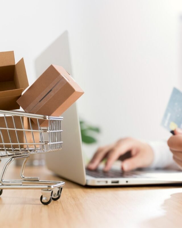 A shopping cart filled with small boxes beside a person using a laptop, holding a credit card, ready to make an online purchase to indicate Cambodia E-commerce Market Trends.