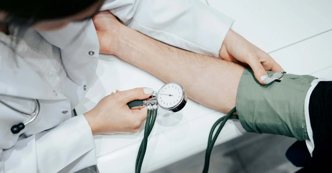 ahmed-6tRWDCpZ1MY-unsplash(1) A healthcare professional measures a patient's blood pressure using a sphygmomanometer on a clinical white surface, Cambodia Healthcare Market Expansion.