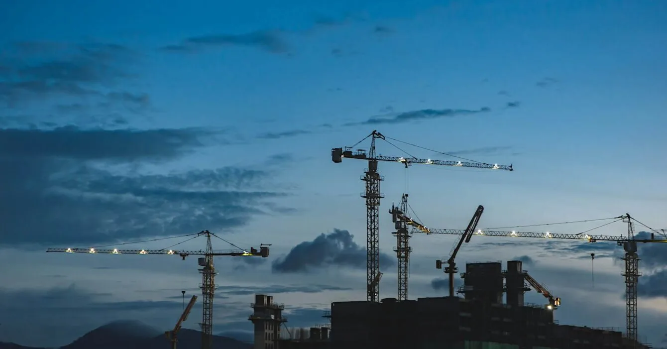 Cambodia Construction Industry Growth: Silhouettes of multiple construction cranes against a twilight sky, with clouds and distant mountains in the background.