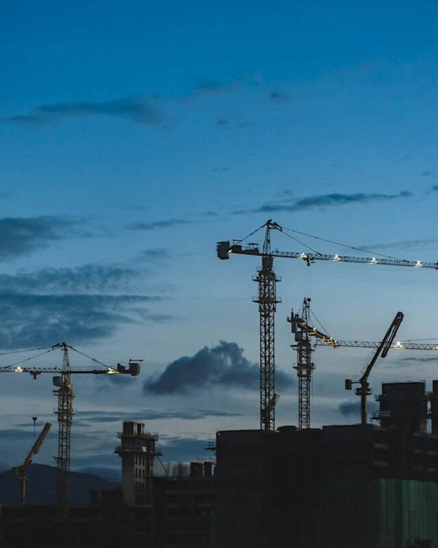 Cambodia Construction Industry Growth: Silhouettes of multiple construction cranes against a twilight sky, with clouds and distant mountains in the background.