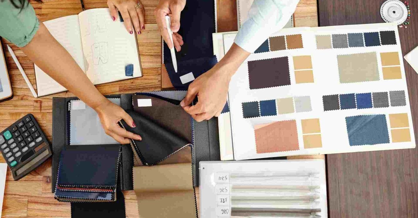 Cambodia Textile Export Strategy: A collaborative workspace featuring hands choosing fabric swatches and reviewing design notes, with tools and sketches scattered on a table.