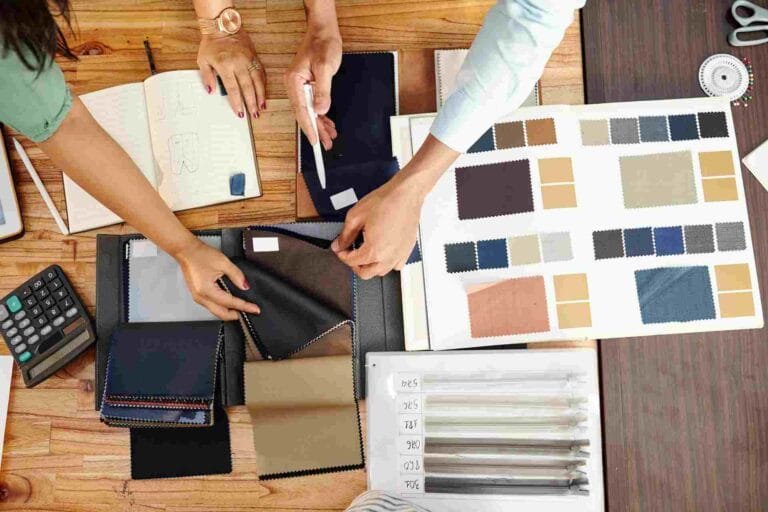Cambodia Textile Export Strategy: A collaborative workspace featuring hands choosing fabric swatches and reviewing design notes, with tools and sketches scattered on a table.