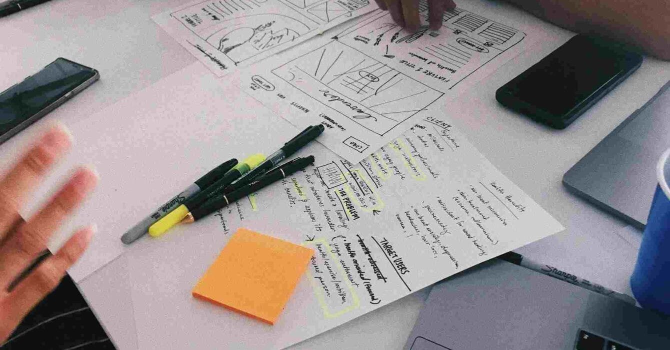 Cambodia Startup Ecosystem Development: Hands and devices around a table with sketches, notes, and stationery, showcasing a collaborative design brainstorming session.