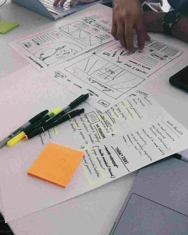 Cambodia Startup Ecosystem Development: Hands and devices around a table with sketches, notes, and stationery, showcasing a collaborative design brainstorming session.