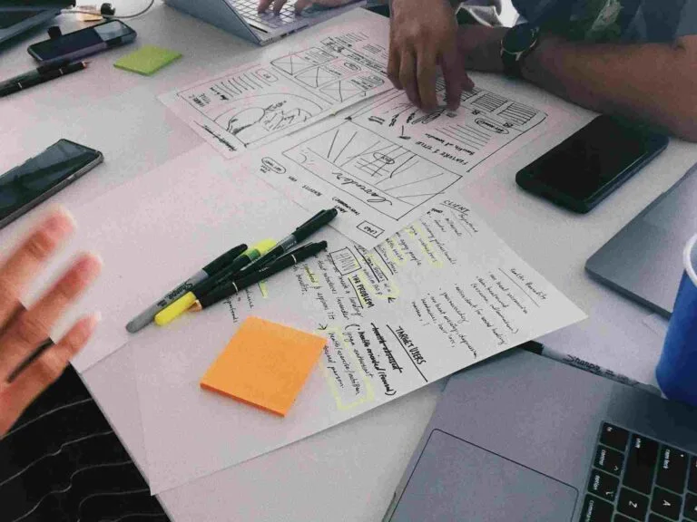 Cambodia Startup Ecosystem Development: Hands and devices around a table with sketches, notes, and stationery, showcasing a collaborative design brainstorming session.