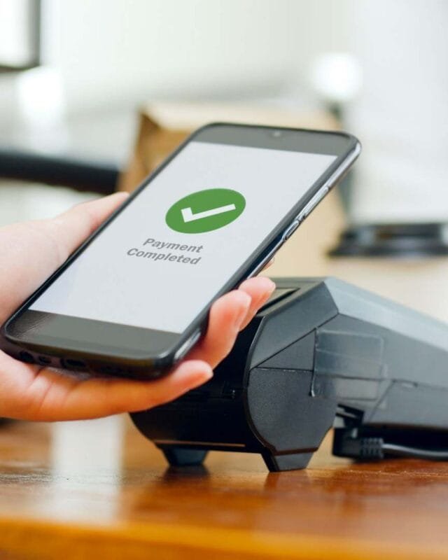 Cambodia QR Code Payment Integration: A person in a yellow sweater uses their smartphone to confirm a completed payment at a contactless terminal on a wooden counter.