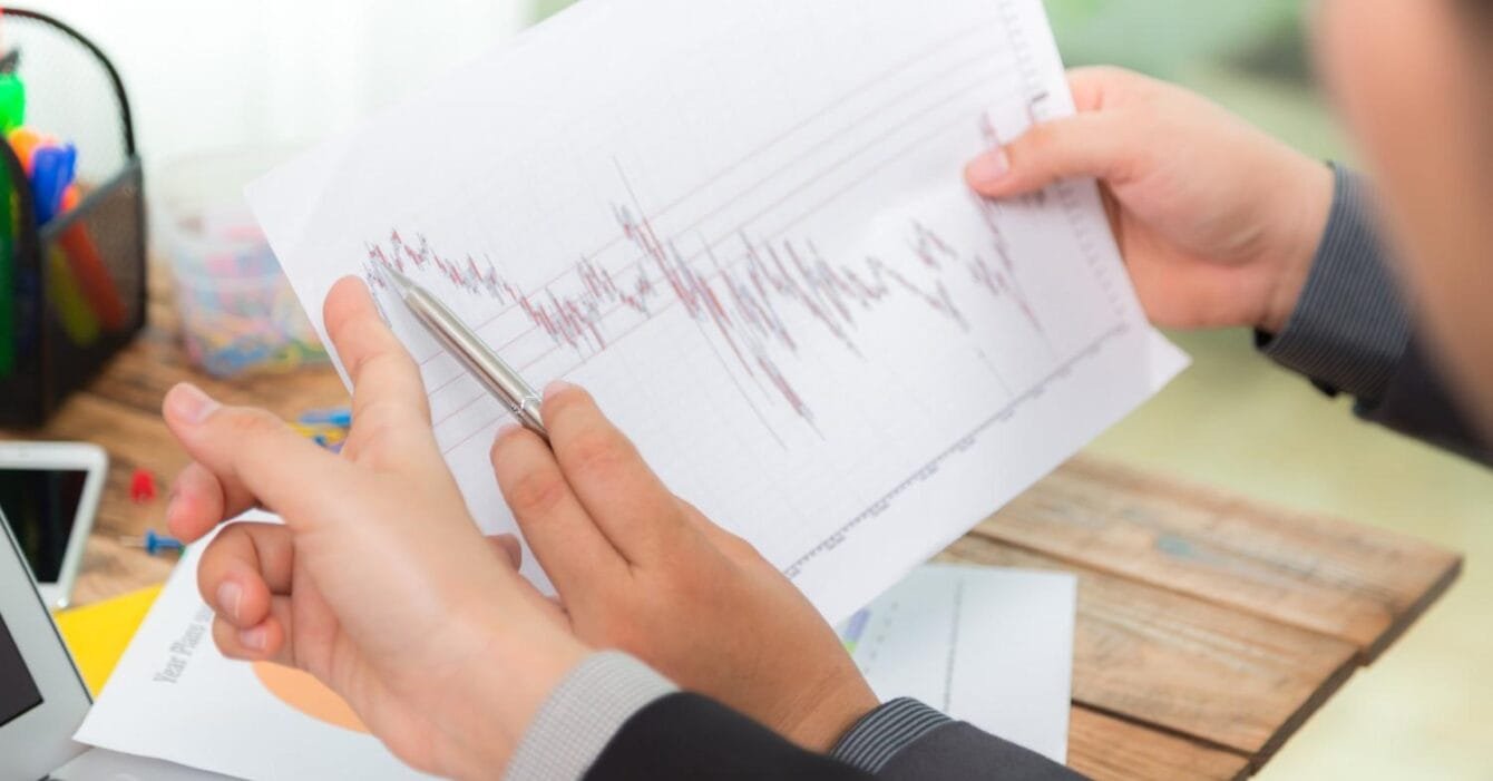 Cambodia GDP Forecast Downgrade: A person in a suit analyzes a financial graph on paper, using a pen to point out trends while seated at a desk with a laptop.