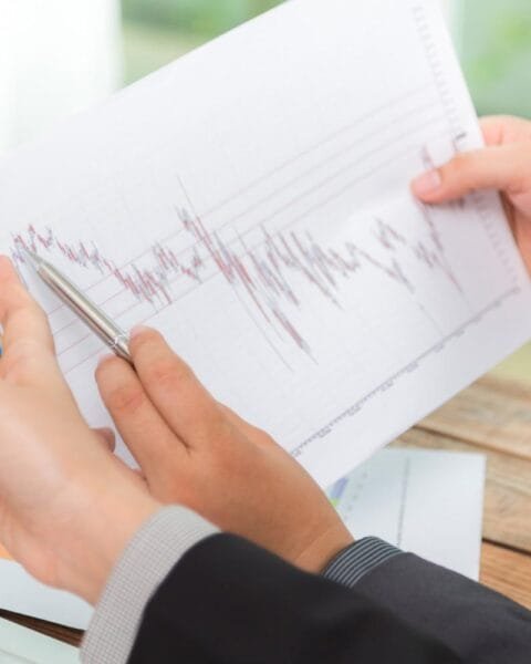 Cambodia GDP Forecast Downgrade: A person in a suit analyzes a financial graph on paper, using a pen to point out trends while seated at a desk with a laptop.