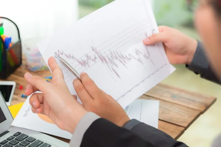 Cambodia GDP Forecast Downgrade: A person in a suit analyzes a financial graph on paper, using a pen to point out trends while seated at a desk with a laptop.