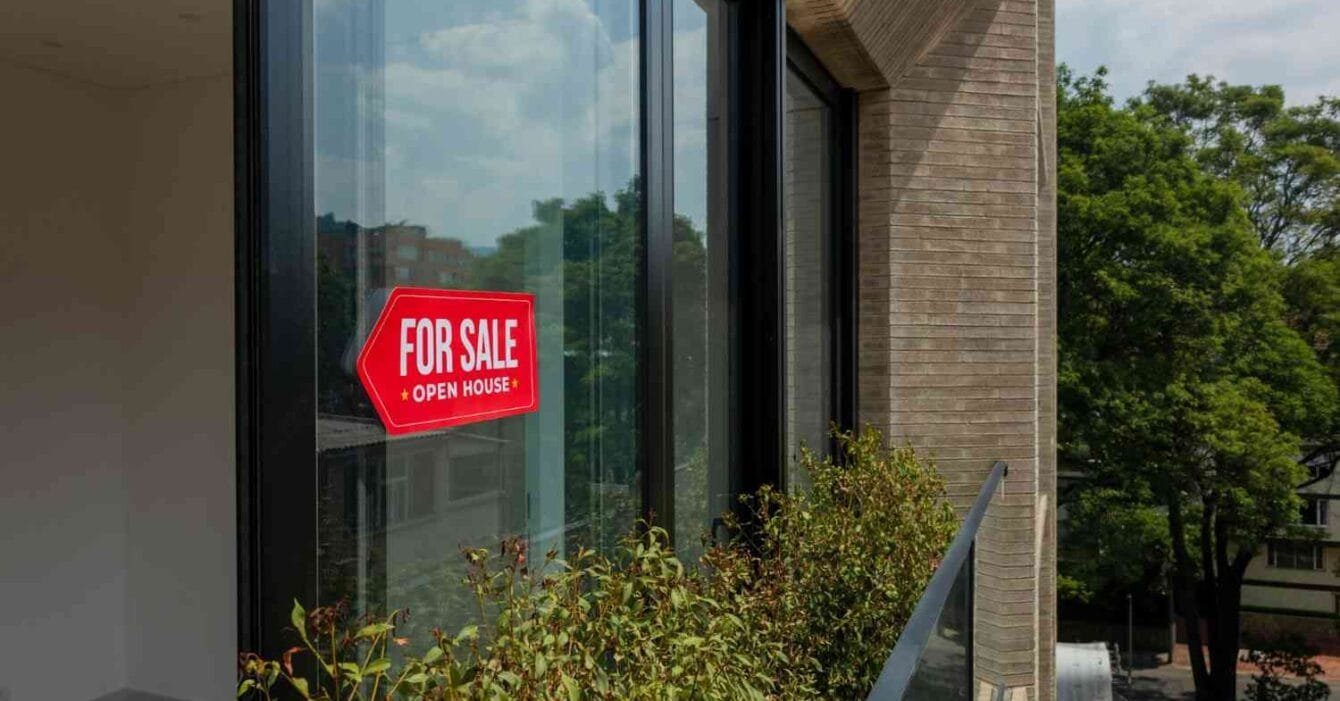 Cambodia Property Market 2025: A bright red "For Sale" sign hangs on a modern window, with lush greenery visible outside and clear skies above.