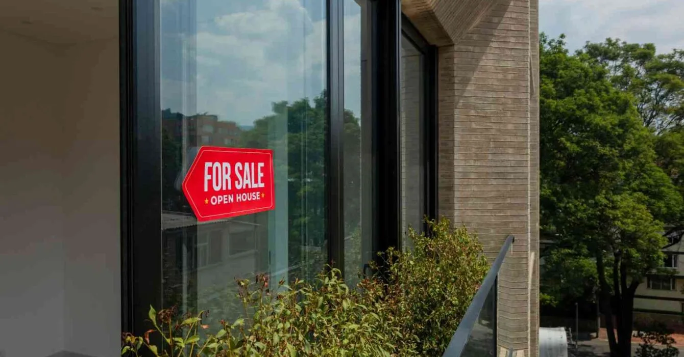 Cambodia Property Market 2025: A bright red "For Sale" sign hangs on a modern window, with lush greenery visible outside and clear skies above.