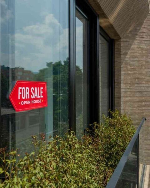 Cambodia Property Market 2025: A bright red "For Sale" sign hangs on a modern window, with lush greenery visible outside and clear skies above.