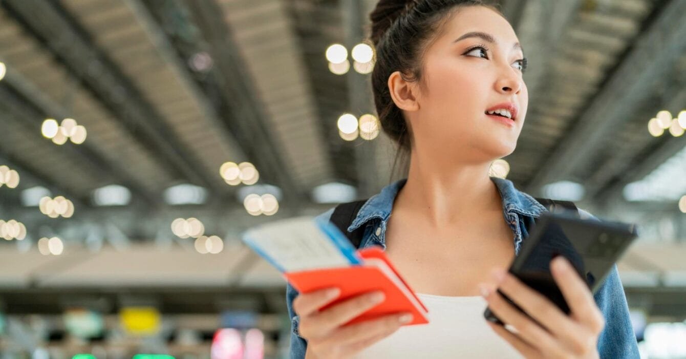 Cambodia Smart Tourism Infrastructure: A woman holds a phone and travel documents in an airport, surrounded by bright lights and bustling activity.