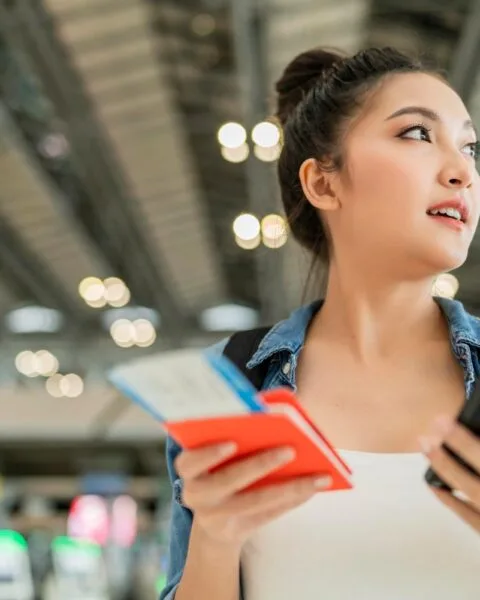 Cambodia Smart Tourism Infrastructure: A woman holds a phone and travel documents in an airport, surrounded by bright lights and bustling activity.