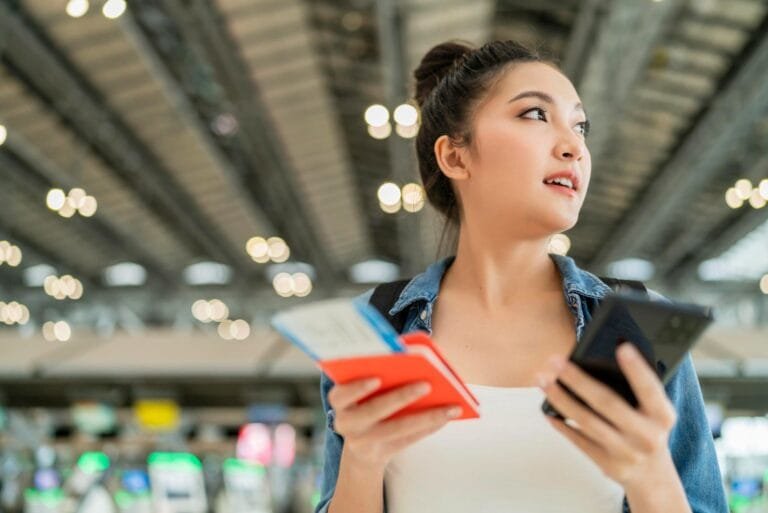 Cambodia Smart Tourism Infrastructure: A woman holds a phone and travel documents in an airport, surrounded by bright lights and bustling activity.