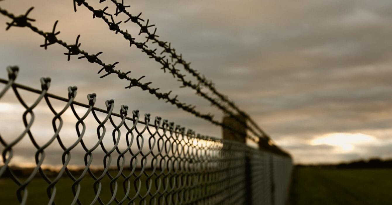 Cambodia Border Tension Effects: Close-up of a barbed wire fence against a cloudy dusk sky, with a blurred foreground and a grassy field in the background.