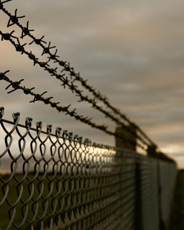 Cambodia Border Tension Effects: Close-up of a barbed wire fence against a cloudy dusk sky, with a blurred foreground and a grassy field in the background.