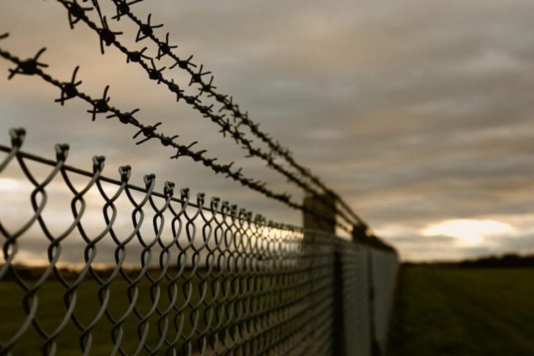Cambodia Border Tension Effects: Close-up of a barbed wire fence against a cloudy dusk sky, with a blurred foreground and a grassy field in the background.