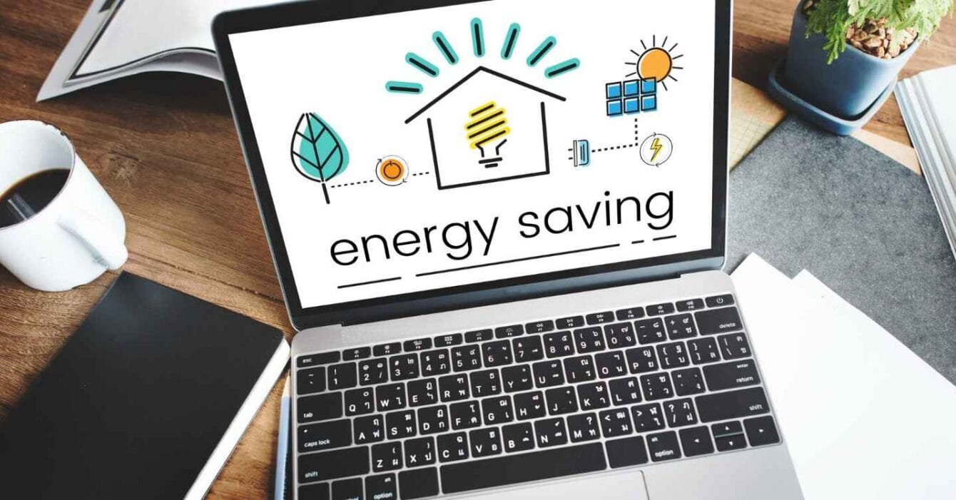 132216 Cambodia Energy Deal Watch: A laptop displays an energy saving graphic with icons of a house, light bulb, sun, and leaf, alongside a cup of coffee and a notebook.