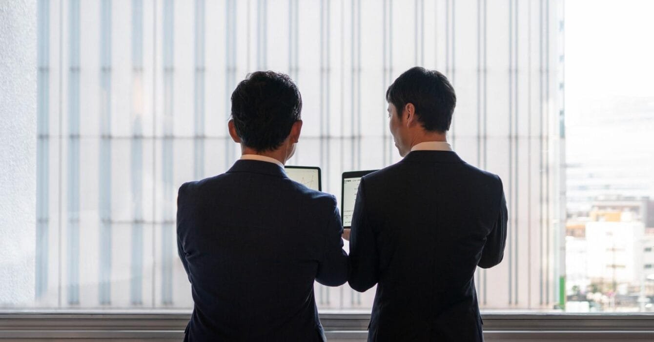 2150103583 Cambodia Border Trade Reopening: Two business professionals in suits, standing by a window, examining information on laptops against a backdrop of a high-rise building.