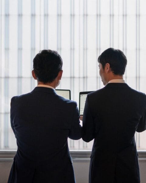 Cambodia Border Trade Reopening: Two business professionals in suits, standing by a window, examining information on laptops against a backdrop of a high-rise building.