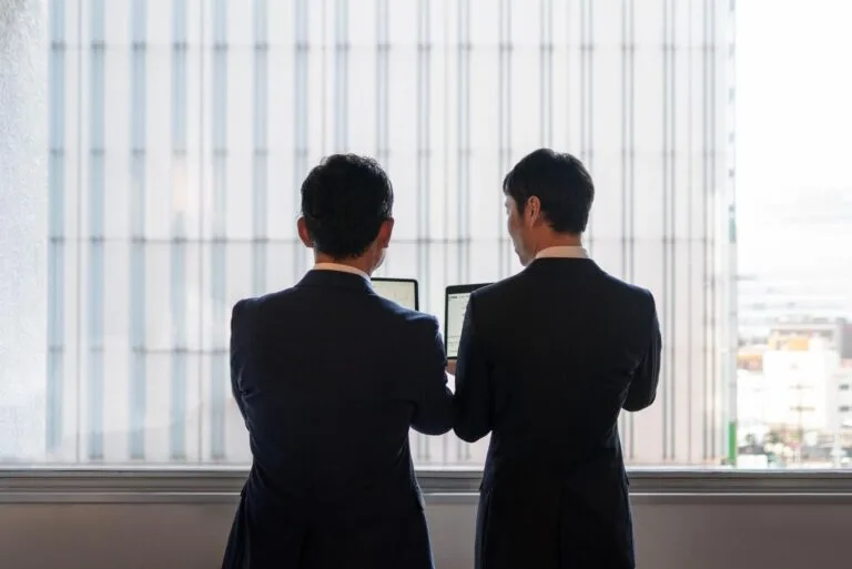 Cambodia Border Trade Reopening: Two business professionals in suits, standing by a window, examining information on laptops against a backdrop of a high-rise building.