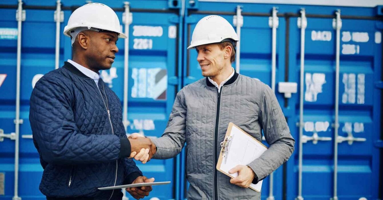 Cambodia's Trade Deal Ambitions: Two construction workers in hard hats shake hands in front of blue shipping containers. One holds a clipboard and tablet.