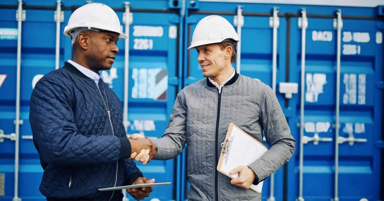 getty-images-LAtqDZZuSQM-unsplash Cambodia's Trade Deal Ambitions: Two construction workers in hard hats shake hands in front of blue shipping containers. One holds a clipboard and tablet.