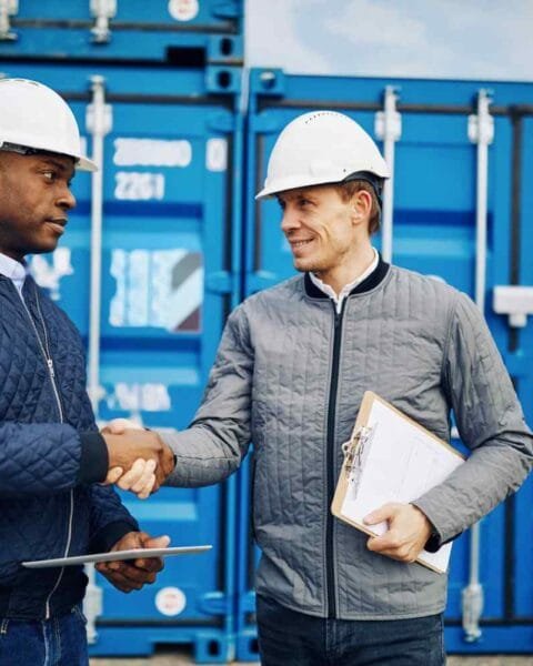 Cambodia's Trade Deal Ambitions: Two construction workers in hard hats shake hands in front of blue shipping containers. One holds a clipboard and tablet.