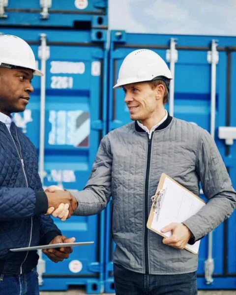Cambodia's Trade Deal Ambitions: Two construction workers in hard hats shake hands in front of blue shipping containers. One holds a clipboard and tablet.