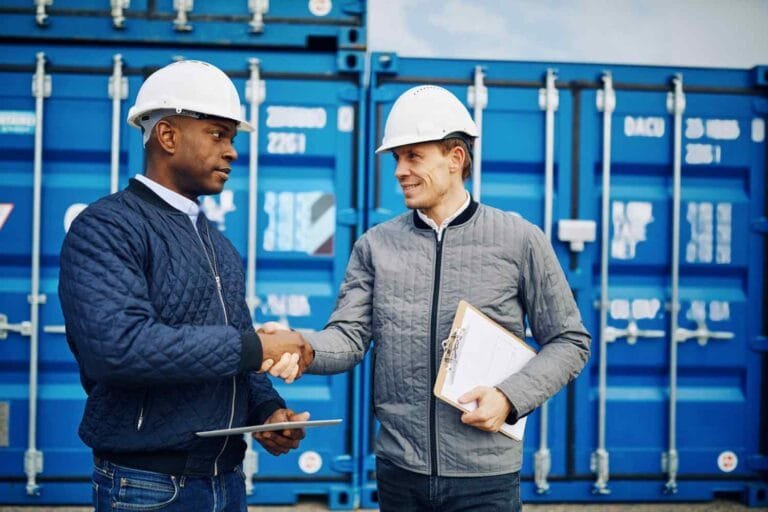 Cambodia's Trade Deal Ambitions: Two construction workers in hard hats shake hands in front of blue shipping containers. One holds a clipboard and tablet.