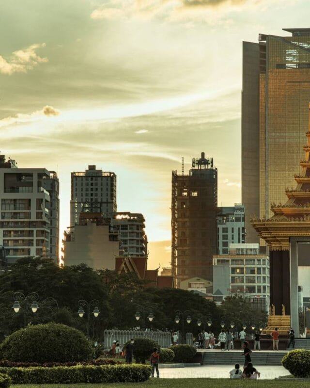 roth-chanvirak-MndC6D5zNDs-unsplash Funan Techo Canal progress: A golden temple with a statue stands amidst modern skyscrapers at sunset, surrounded by people and lush greenery in the foreground.