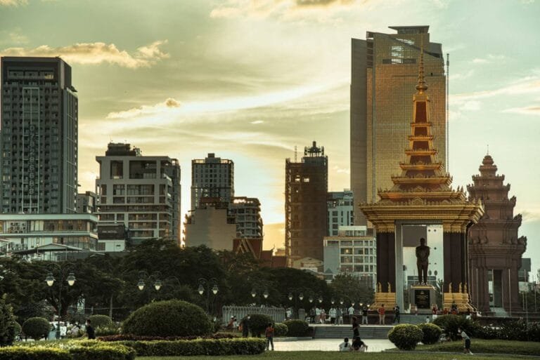 Funan Techo Canal progress: A golden temple with a statue stands amidst modern skyscrapers at sunset, surrounded by people and lush greenery in the foreground.